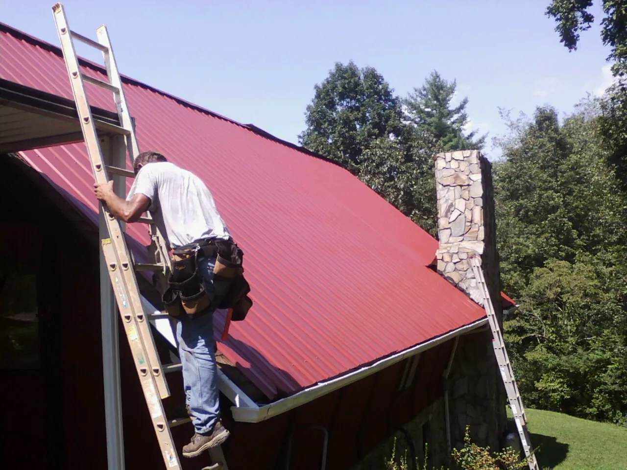 Roofer on a ladder beside a red metal roof and stone chimney — Soffit & Fascia Repair by Roofing Brothers in Cottageville